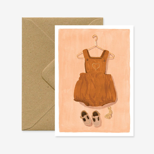 A baby greeting card with a illustration of a brown baby romper suit and shoes on a pink background, accompanied by a brown envelope.