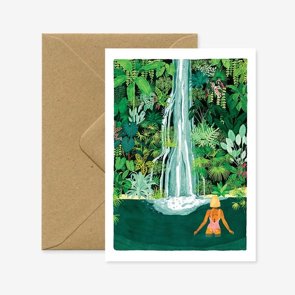 A greeting card with an illustration of a person standing near a waterfall surrounded by greenery. The card is accompanied by a brown envelope.