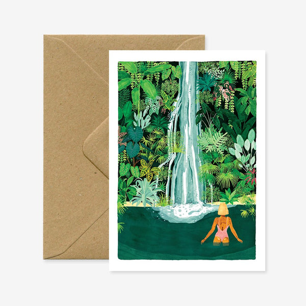 A greeting card with an illustration of a person standing near a waterfall surrounded by greenery. The card is accompanied by a brown envelope.