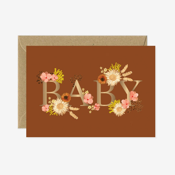 A greeting card with the word 'BABY' written in gold foil, surrounded by floral designs, in an brown envelope.
