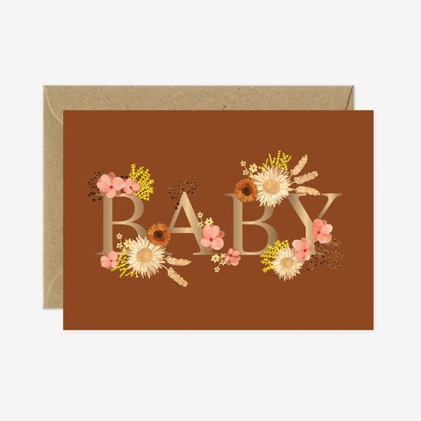 A greeting card with the word 'BABY' written in gold foil, surrounded by floral designs, in an brown envelope.