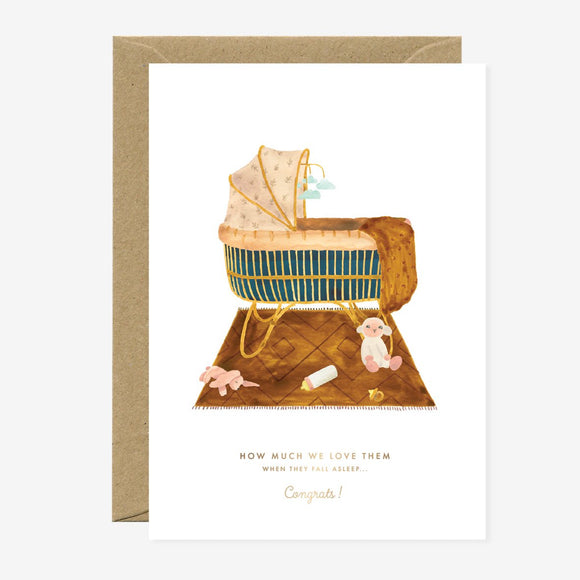 A greeting card depicting an illustration of a baby in a bassinet with gold foil details, accompanied by a teddy bear and a rattle, with a brown envelope.