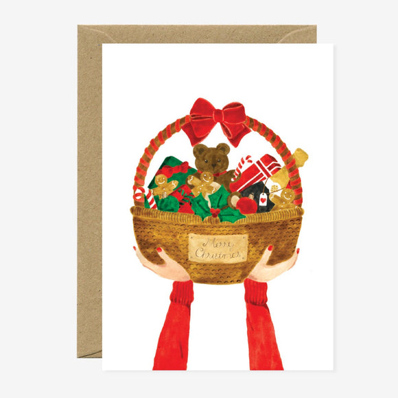 A Christmas themed greeting card depicting a basket with gifts and a teddy bear, held by a pair of hands, with a red bow at the top and the text 'Merry Christmas' on the front.