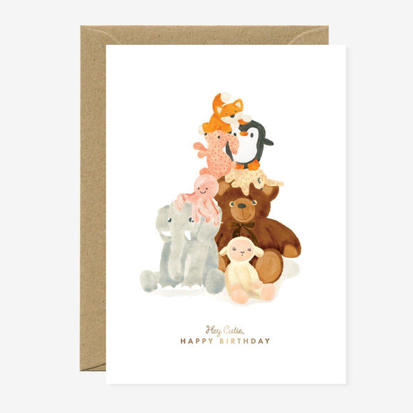 A birthday greeting card with a gold foil detailed illustration of various stuffed animals such as a bear and a penguin, with a 'Happy Birthday' text at the bottom. There is a brown envelope behind the card.