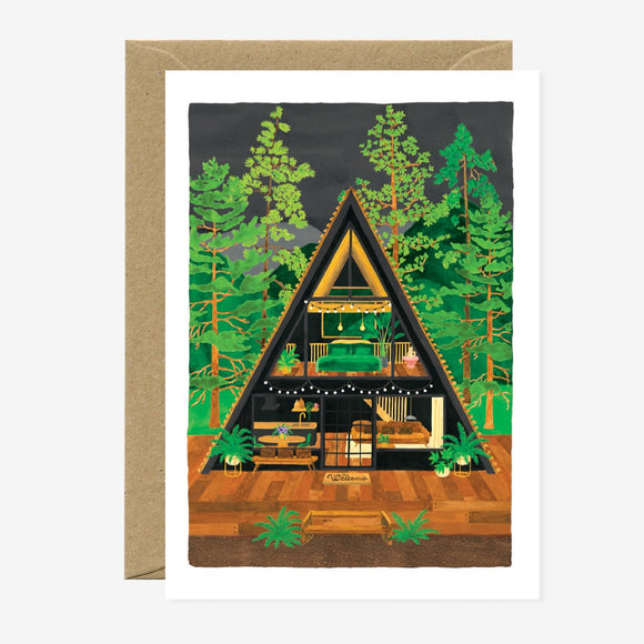A greeting card depicting a triangular cabin in a forest setting, with gold foil details and a brown envelope.