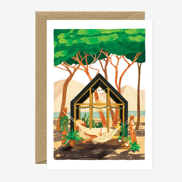 A greeting card featuring an illustration of a person relaxing in a hammock by a beach, with trees and a wooden structure in the background. The card includes gold foil details and comes with a brown envelope.