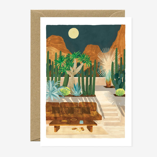 A greeting card with an illustration of a desert landscape, featuring plants, mountains, and a clear sky, with gold foil details. A brown envelope is included.