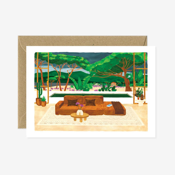 A greeting card featuring an illustration of a cozy living room with a view of a lush outdoor landscape. The card is accompanied by a brown envelope.