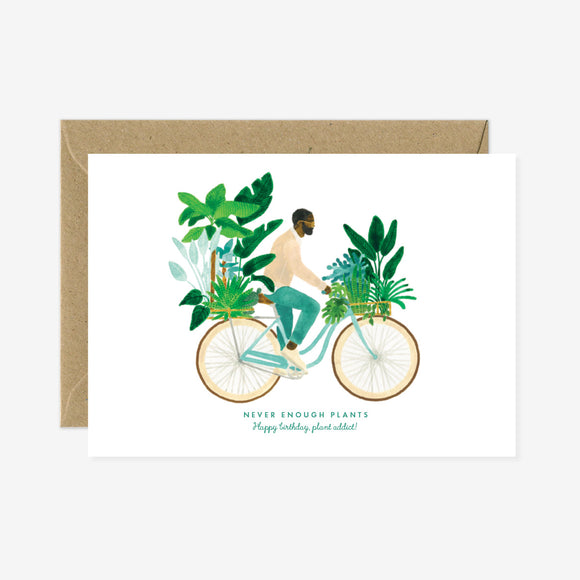A birthday greeting card with a illustration of a person on a bicycle surrounded by plants, with a gold foil detail and a brown envelope.