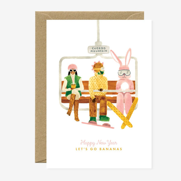 A Christmas greeting card featuring an illustration of a person, a reindeer, and a rabbit sitting on a bench with ski equipment, accompanied by the text 'Happy New Year - LET'S GO BANANAS' and a recycled brown envelope.