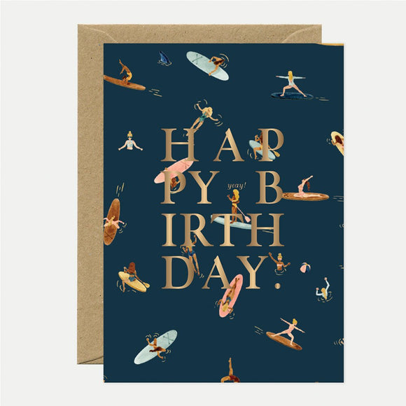 A birthday greeting card with the text 'HAPPY BIRTHDAY' in gold foil, surrounded by illustrations of people paddle-boarding in a navy background. The card is accompanied by a brown envelope.