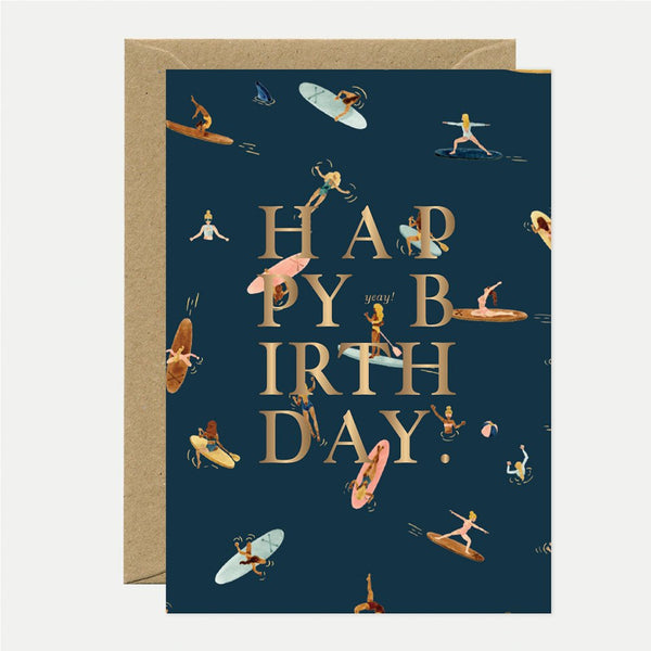 A birthday greeting card with the text 'HAPPY BIRTHDAY' in gold foil, surrounded by illustrations of people paddle-boarding in a navy background. The card is accompanied by a brown envelope.