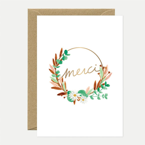 A white greeting card with 'Merci' written in gold foil, surrounded by a floral wreath design in gold, green, and brown colors, accompanied by a brown envelope.