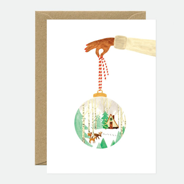 A Christmas greeting card featuring a hand holding a round ornament with a forest and snowscape scene inside, accompanied by a brown envelope.