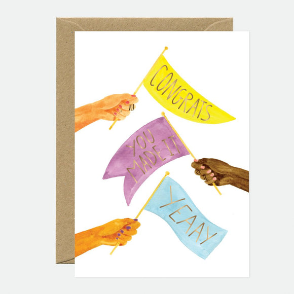 A greeting card with a design of four hands holding flags with messages 'CONGRATS', 'SO Proud', and 'YEAH'. The card has a gold foil detail and is placed next to a brown envelope.