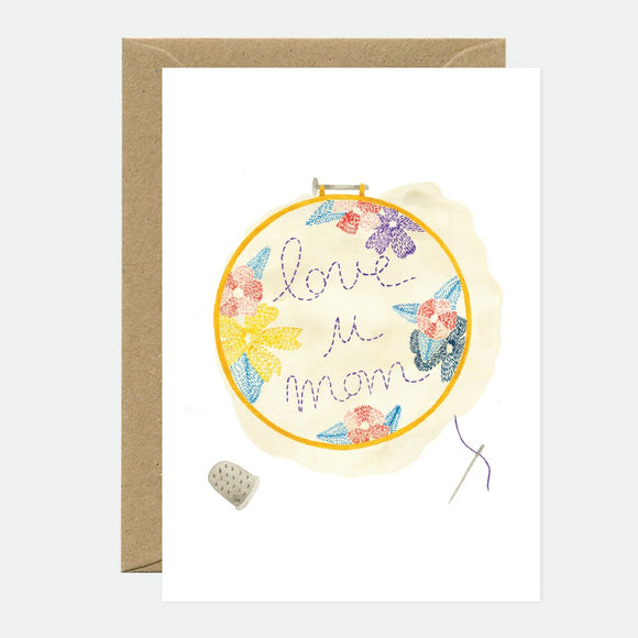 A Mother's Day greeting card with floral embroidery design and the text 'love u mom' in a hoop, accompanied by a brown envelope.