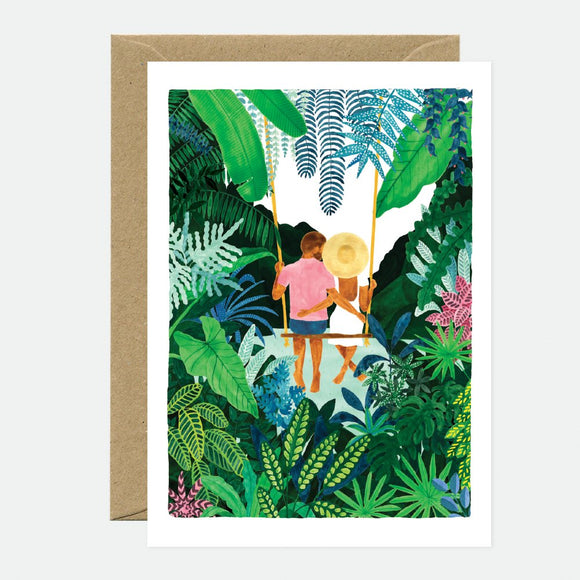 A greeting card with an illustration of two people on swings surrounded by lush greenery. The card is accompanied by a brown envelope.