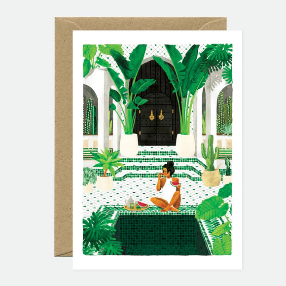 A greeting card depicting a scene with greenery and a figure, with gold foil details, accompanied by a brown envelope.
