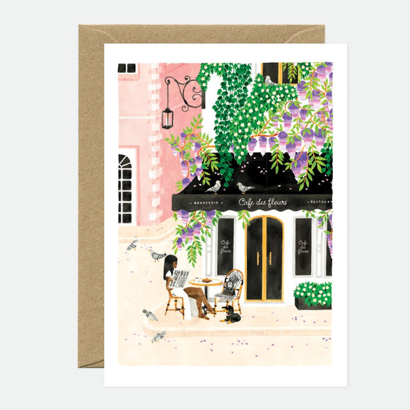 Illustrated greeting card depicting a scene with a cafe, trees, and a building facade, accompanied by a brown envelope.