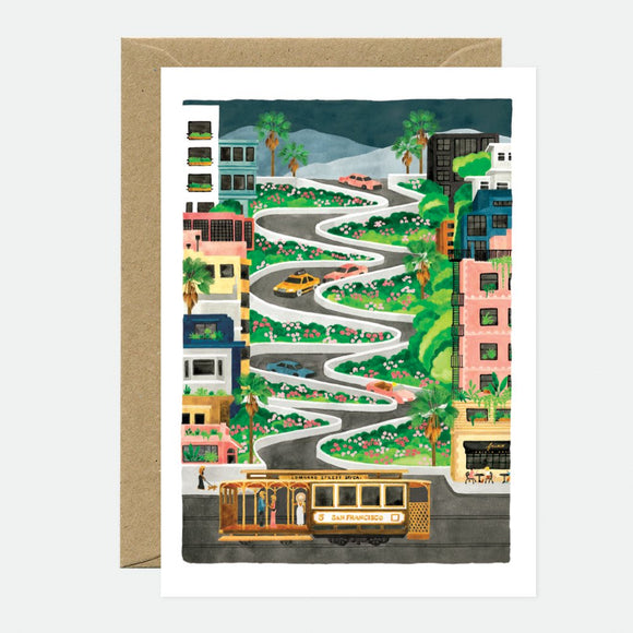 A greeting card featuring an illustration of a winding road with houses and a cable car, suggestive of a travel landscape. The card is accompanied by a brown envelope.