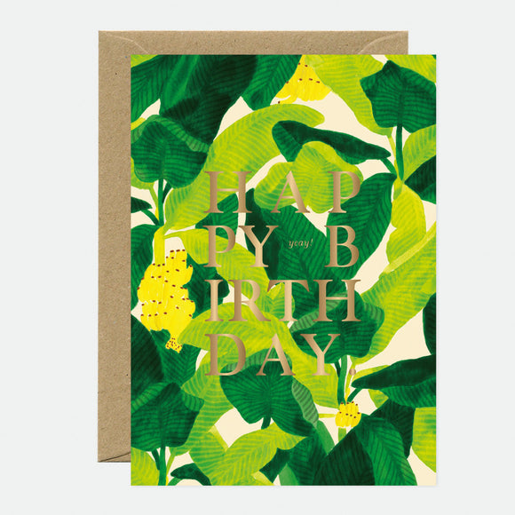 A birthday greeting card with gold foil text that reads 'HAPPY BIRTHDAY', featuring a green leafy background, accompanied by a brown envelope.