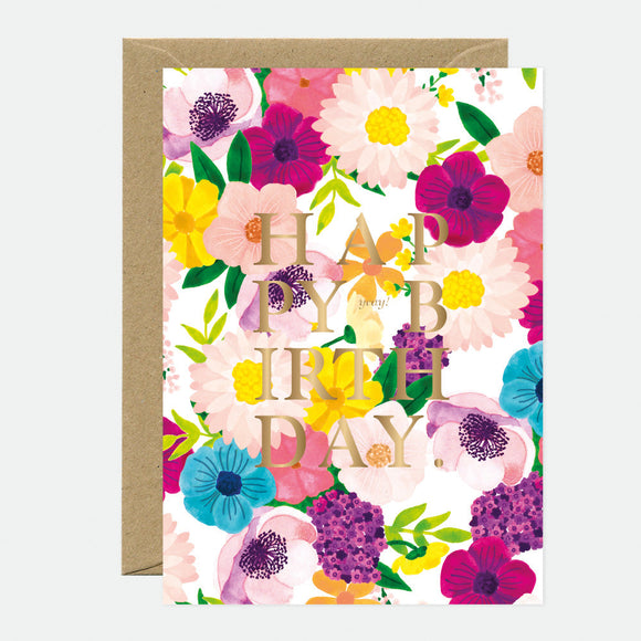 A birthday greeting card with a gold foil text reading 'HAPPY BIRTHDAY' surrounded by a floral pattern in shades of pink, purple, yellow, and green, accompanied by a brown envelope.