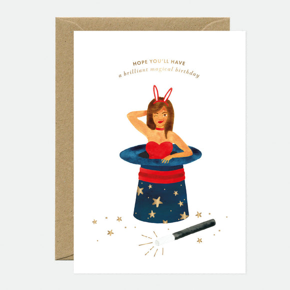 A greeting card with a illustration of a person coming out of a magician's hat, with gold foil details and stars, accompanied by a brown envelope.