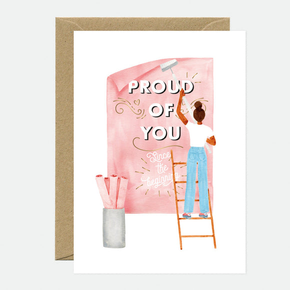 A greeting card with a gold foil inscription saying 'Proud of You' accompanied by an illustration of a person on a ladder writing on a pink board, with a brown envelope in the background.