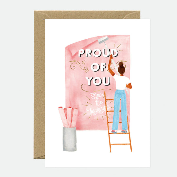 A greeting card with a gold foil inscription saying 'Proud of You' accompanied by an illustration of a person on a ladder writing on a pink board, with a brown envelope in the background.