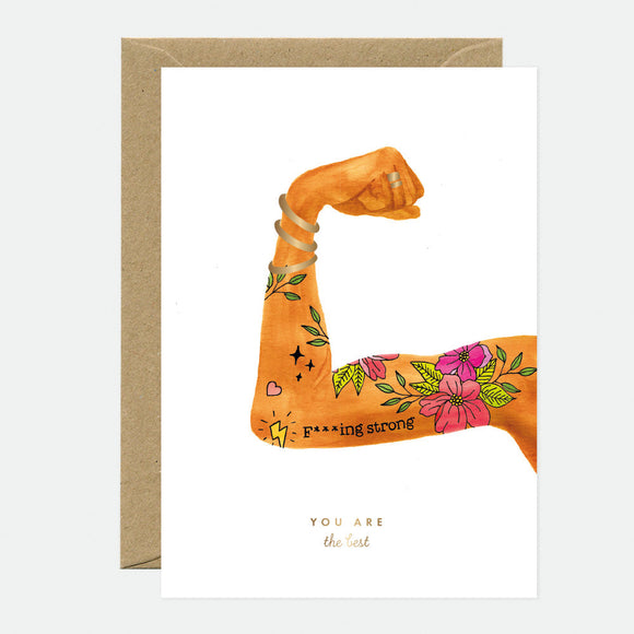 A greeting card with an illustration of a person's flexed bicep with floral tattoos and the text 'F***ing strong - YOU ARE the best' on it. The card is accompanied by a brown envelope.