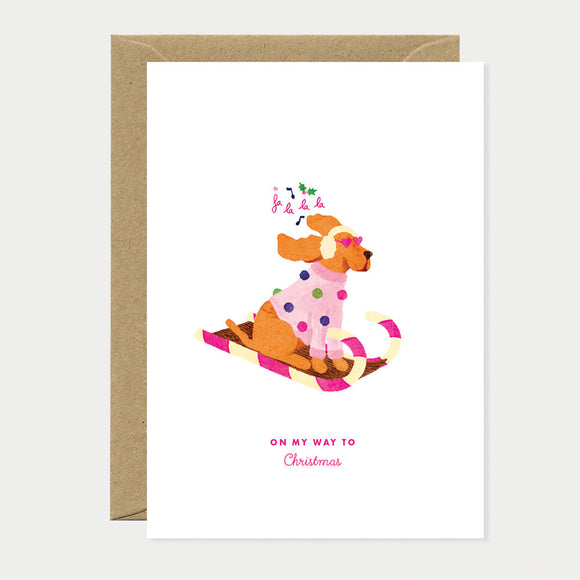 Greeting card with a festive dog riding a candy-cane sled, wearing earmuffs and scarf, symbolizing playful Christmas spirit.