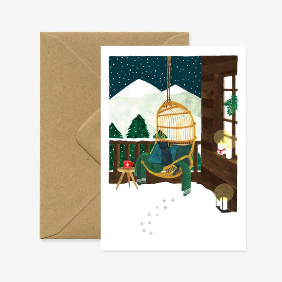 A Christmas greeting card depicting a cozy chalet with a person sitting in a chair by a window, looking out at a snowy landscape, accompanied by a brown envelope.