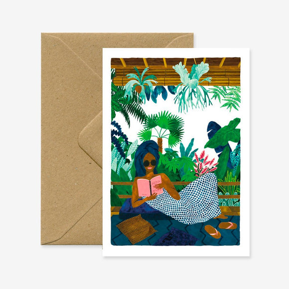 A greeting card with an illustration of a person reading a book in a tropical setting, accompanied by a brown envelope.