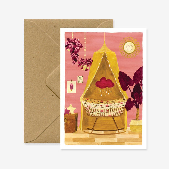 A greeting card with a illustration of a baby crib and decorative elements like plants and a sun in the background. The card is displayed with a brown envelope.