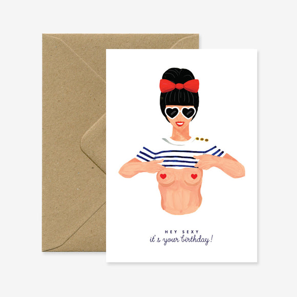 A greeting card with a illustration of a stylized woman with a red bow and sunglasses, holding out a t-shirt and text that reads 'Hey Sexy ! It's your birthday!'. There is a brown envelope behind the card.