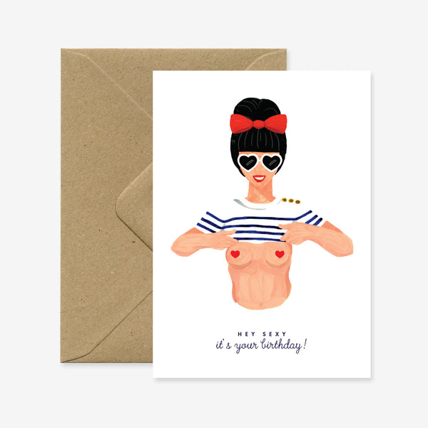 A greeting card with a illustration of a stylized woman with a red bow and sunglasses, holding out a t-shirt and text that reads 'Hey Sexy ! It's your birthday!'. There is a brown envelope behind the card.