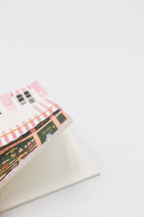 Open dotted notebook with illustrated pink bakery storefront cover, showing clean interior pages on a white background.