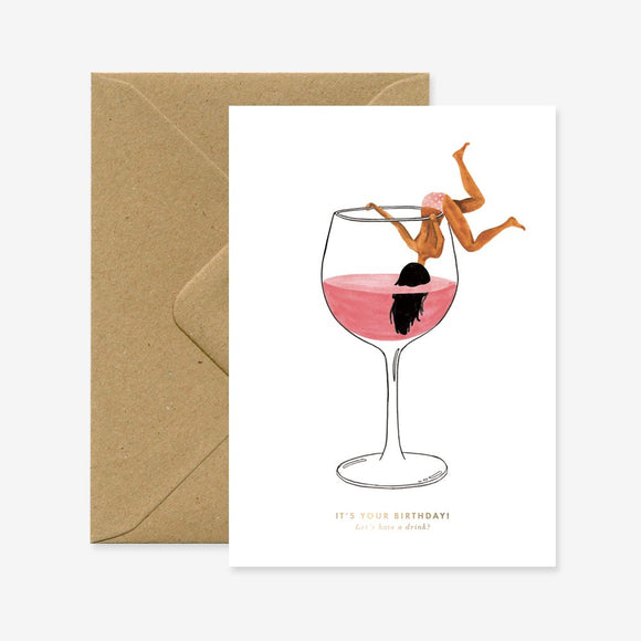 A birthday greeting card with a illustration of a woman inside a glass of pink liquid, accompanied by a brown envelope.