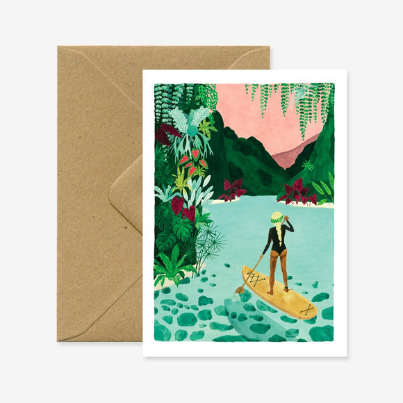 A travel greeting card with a illustration of a person paddle-boarding in a tropical setting. Accompanied by a brown envelope.
