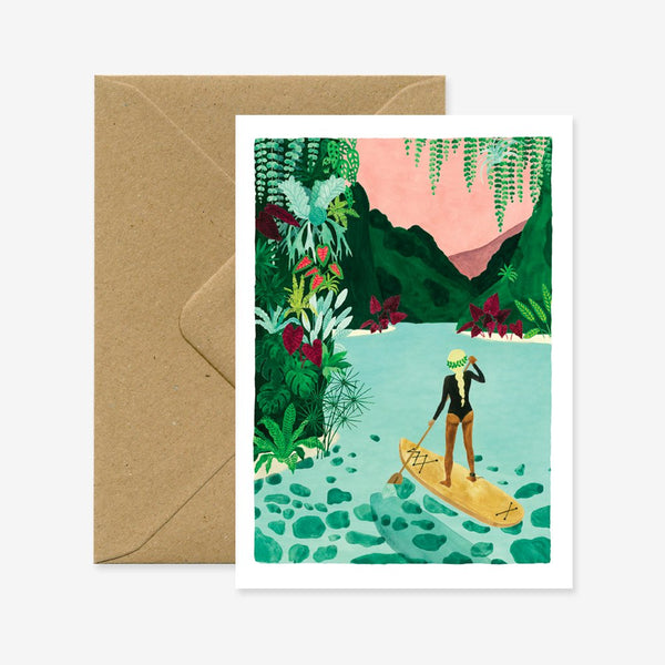 A travel greeting card with a illustration of a person paddle-boarding in a tropical setting. Accompanied by a brown envelope.