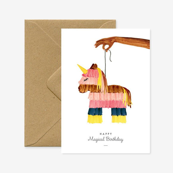 A birthday greeting card with a colorful piñata designed to look like a unicorn, held by a hand, with a brown envelope in the background. The text on the card reads 'Happy Magical Birthday'.