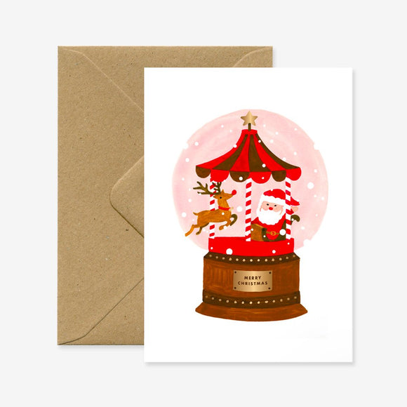 A Christmas greeting card featuring an illustration of Santa Claus inside a snow globe with a reindeer, trees, and the text 'Merry Christmas' on the front, accompanied by a brown envelope.
