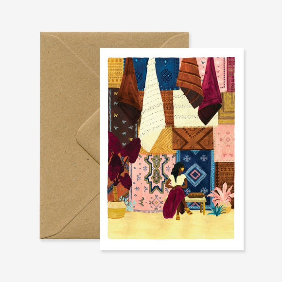 A greeting card with an illustration of a travel landscape, showing various fabrics and a market scene, accompanied by a brown envelope.