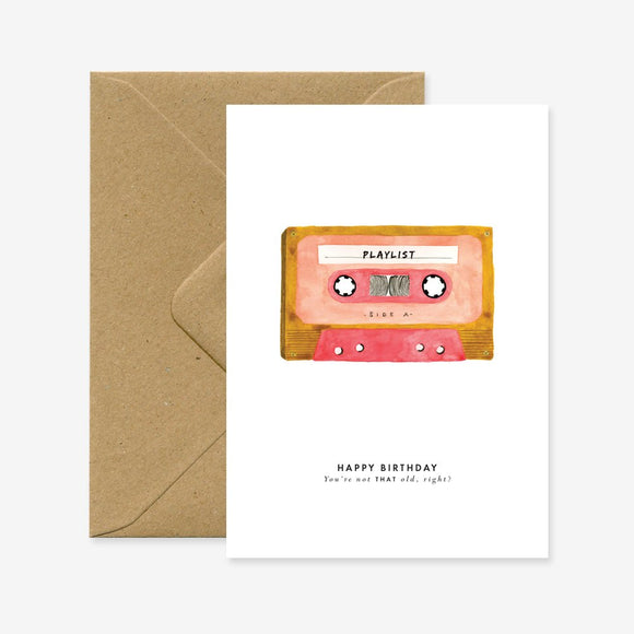 A birthday greeting card with a illustration of a cassette tape on the front, accompanied by a brown envelope. The text on the card reads 'Happy Birthday - You're one year cooler, right?'.
