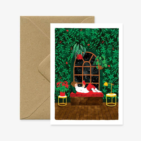 A Christmas themed greeting card depicting a cartoon illustration of Santa Claus sitting in a chair with a Christmas tree and gifts in the background, accompanied by a brown envelope.