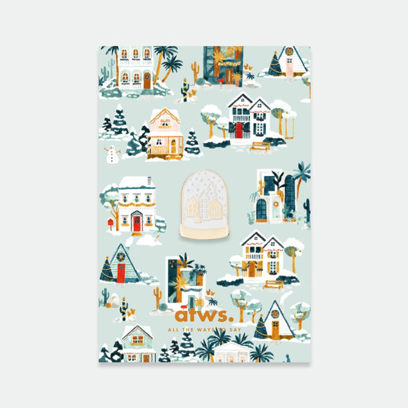 A collection of enamel pins with a winter theme, including houses and a snow globe, on a brand card.