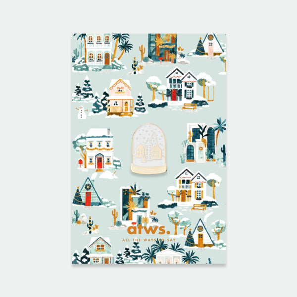 A collection of enamel pins with a winter theme, including houses and a snow globe, on a brand card.