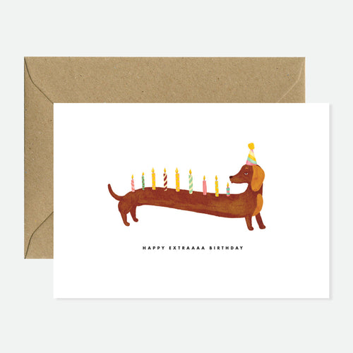 A greeting card with an illustration of a dachshund wearing a party hat with candles, indicating it is for a birthday. The card is displayed with a natural white envelope.