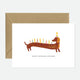 A greeting card with an illustration of a dachshund wearing a party hat with candles, indicating it is for a birthday. The card is displayed with a natural white envelope.