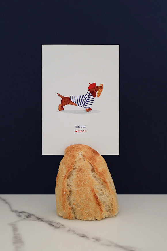 All The Ways To Say Oui Oui Merci card – dachshund in striped shirt holding baguette, styled with bread and marble backdrop